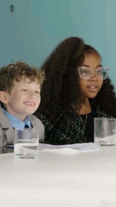 Two children in formal attire sit at a conference table, with glasses of water, in a modern meeting room.