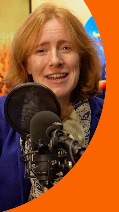 Dr Joelle Grogan sits behind a microphone. She has red hair, and is wearing a blue velvet jacket with a patterned brown and gold scarf.