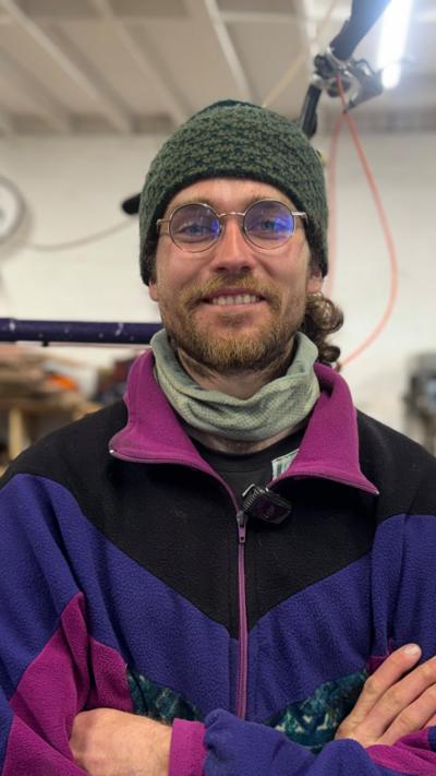 A man wearing a green hat and glasses looks into the camera smiling. He is wearing a purple coat with pink and black trim, and a light green scarf. He has a dark beard and has his arms folded.