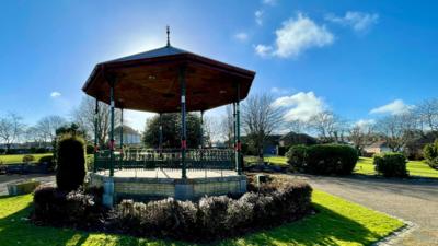 A bandstand in a park surrounded by green shrubs with blue sky and sunshine above