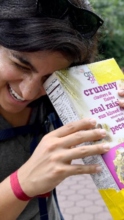 A woman smiling with the corner of a cereal box places against her left eye.