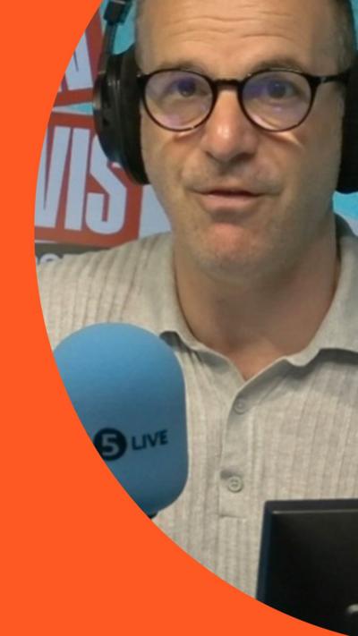 Martin Lewis, wearing headphones and brown glasses, speaks into a 5 Live blue mic