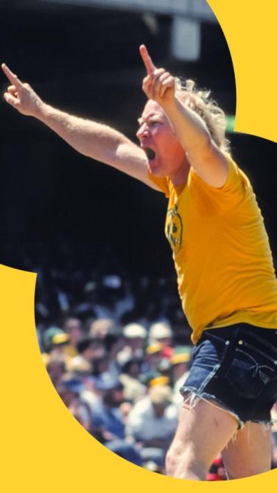 An old man in a yellow shirt and denim shorts raises his index fingers in the air at a sporting event.