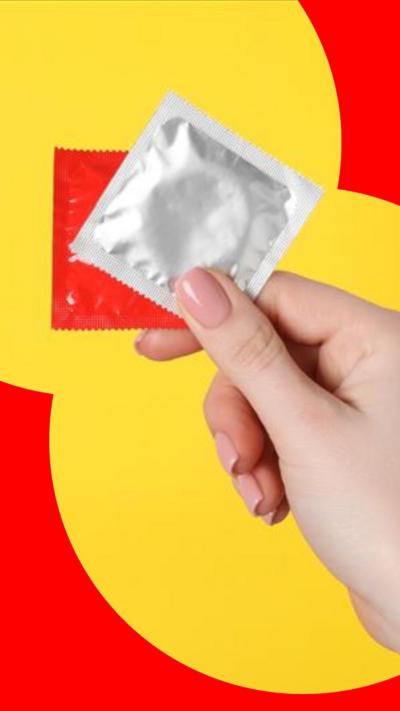 A woman's hand holding up a red and a silver condom packet, in front of a yellow background.