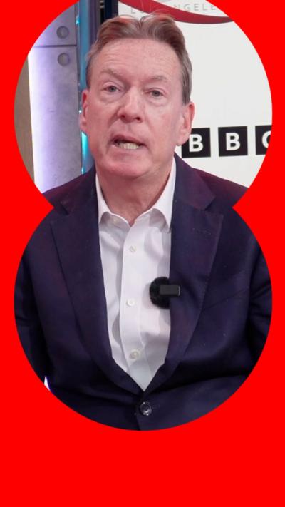Frank Gardner is sat down and wearing a navy suit with a white shirt. Behind him is a screen with the BBC logo on.