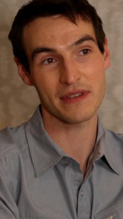 Éanna Hardwicke has short dark hair and is wearing a blue shirt. He has his head tilted and is looking away from the camera.