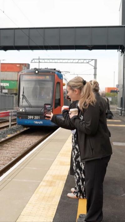 People stand on a tram platform taking photos of a blue red and orange tram