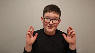 A boy wearing glasses looks at the camera, crossing his fingers