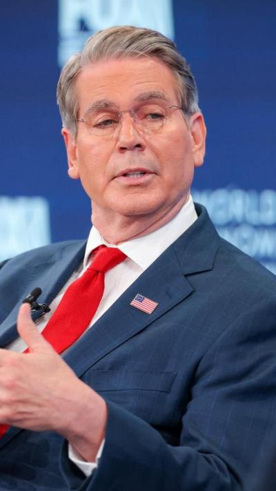 Scott Bessent sits down and talks at the World Economic Forum. He wears a red tie and blue suit with an American flag pin on his lapel.