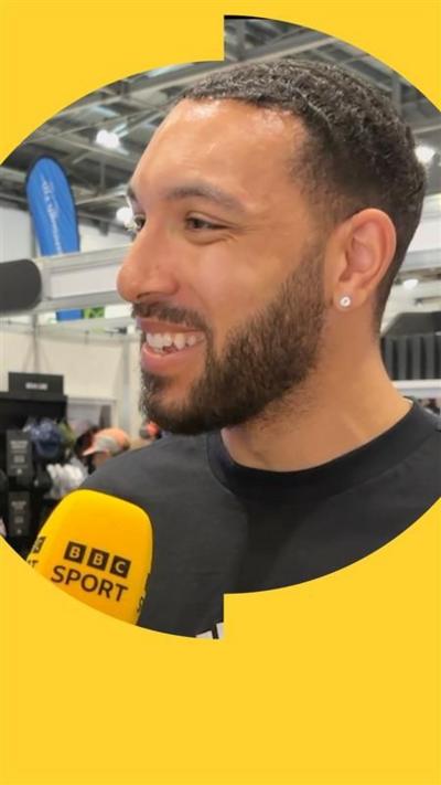 A man smiling, wearing a black t-shirt, with a yellow microphone in front of him =,