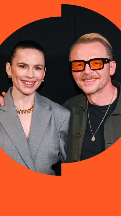 Hayley Atwell in a grey suit and Simon Pegg in a green shirt smile with their arms around each other