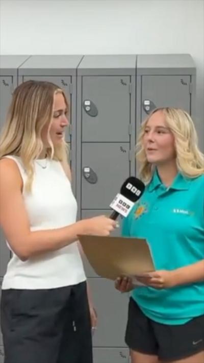 BBC reporter Lucy Woodham talking to a blonde-haired student in a turquoise polo shirt. She is holding an envelope of results