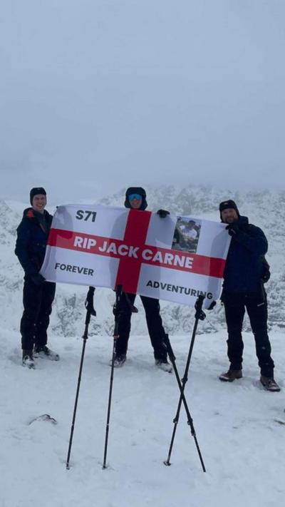 Matty Belcher, Brandan Smith and Jack's dad, Rich, they are all holding the England flag that has 'rip Jack Carne' in capitals on it and also 'forever adventuring' as well. They are standing in front of snowy white mountains.