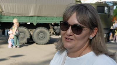 A woman in sunglasses stands in front of an army vehicle