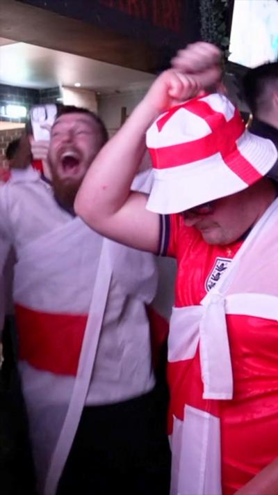 England football fans celebrate in a pub, one is wearing at St George's cross red and white hat and a red football shirt with the Three Lions on, another one has an England flag round his shoulders. 