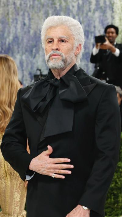 Singer Bad Bunny wears a grey hair wig and facial hair along with a black suit with a large bow around the neck.