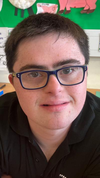 Ben has black hair, black glasses and a black shirt on. He sits in a classroom in front of a noticeboard.