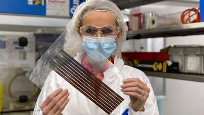 Lara Lewington holds printed solar cells. She is wearing a hair net, goggles and a face mask to protect the electronics