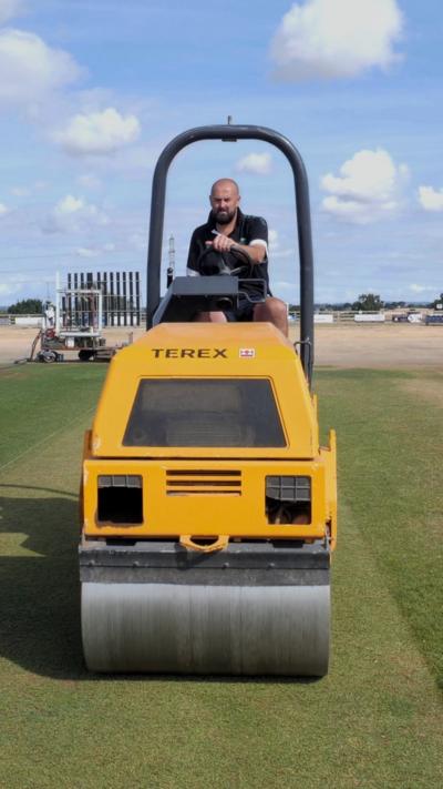 A man in a black top driving a yellow roller on a cricket pitch