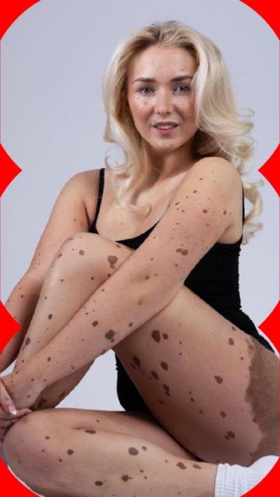 A woman is sitting cross-legged on the floor with a white background. She is smiling at the camera.