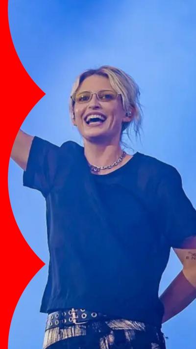 Emily Armstrong smiling with her right arm raised to the sky. She's wearing a black t-shirt and clear glasses.