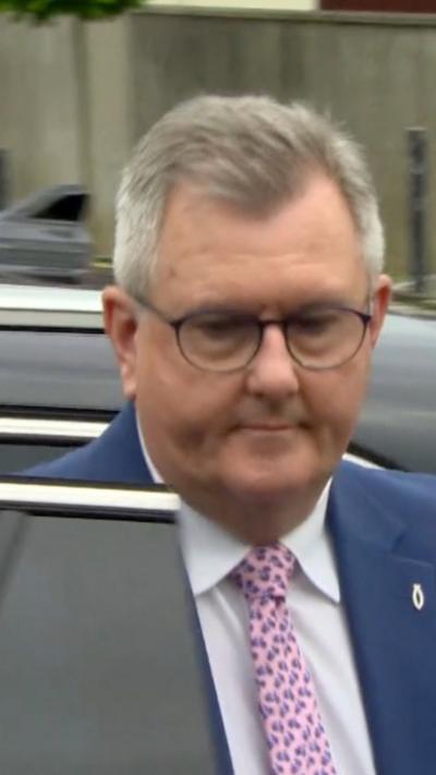 Jeffrey Donaldson is walking away from a black car. He is a man with short grey hair, wearing a dark blue suit, white shirt and pink tie.