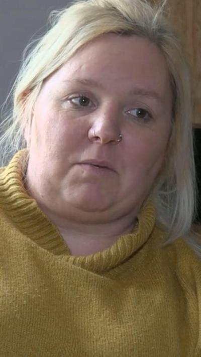 A blond haired woman with a nose ring and brown eyes. She is wearing a yellow jumper. 