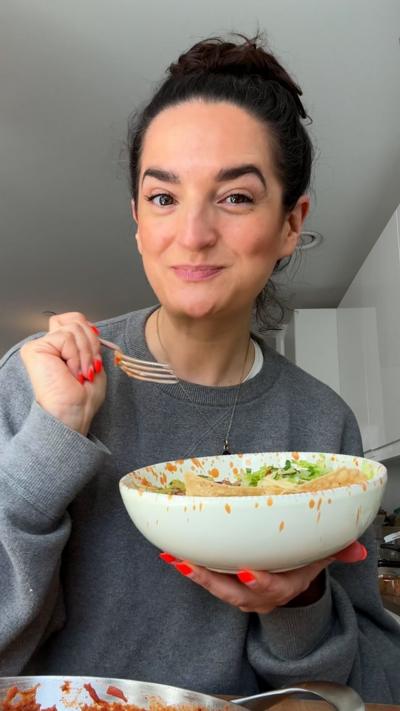 A woman wearing a grey jumper smiles at the camera while holding a bowl of food with her left hand and a fork with her right.