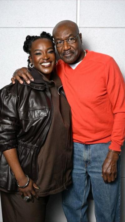 Wunmi Mosaku and Delroy Lindo stand close together for a picture. Delroy, in an orange top, has his arm around Wunmi's shoulder. She wears a brown jacket