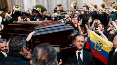 Trombones play as Colon's coffin exits the cathedral.