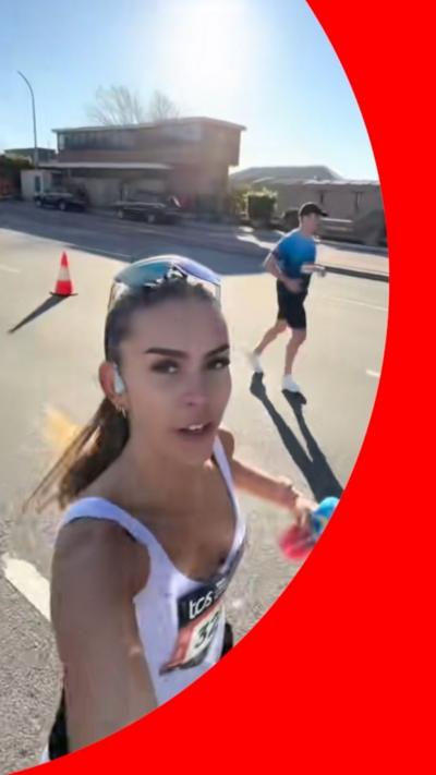 A woman with long brown hair wearing sports kit films herself while running along a road