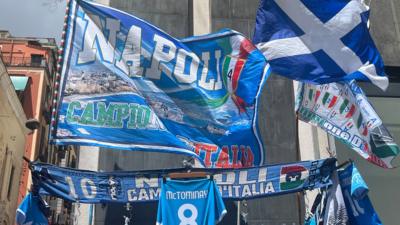 A picture of a blue Napoli flag, a Napoli scarf and a Scotland flag flying in the wind at a vendor's stall in Napoli. A blue football shirt with the name McTominay and the number 8 hangs underneath.