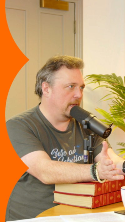 A man is sat at a table with a black mic in front of him that's resting on two red books.