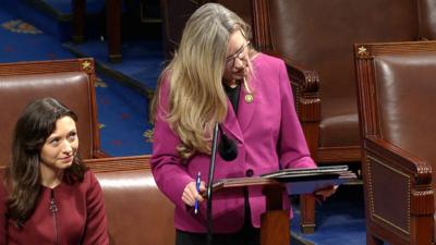 Congresswoman Jennifer Wexton stands in front of a podium while looking at her iPad. She has a pen in her hand. To her right, there is a female staring at her.