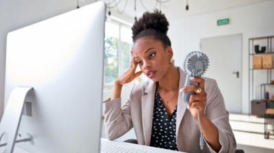 A woman sat in front of a computer screen, holding a mini fan, looking tired