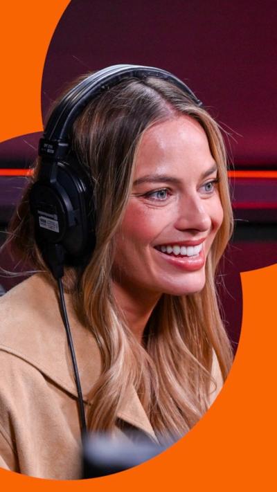 Margot Robbie smiled. She has long brown hair and a beige jacket, with headphones on her head.