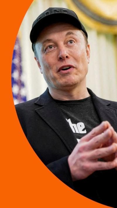 Elon Musk, wearing black, with hands together, fingertips touching, and a baseball cap.