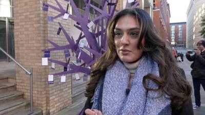 A woman with long brown hair and a purple scarf in front of a purple metal tree sculpture