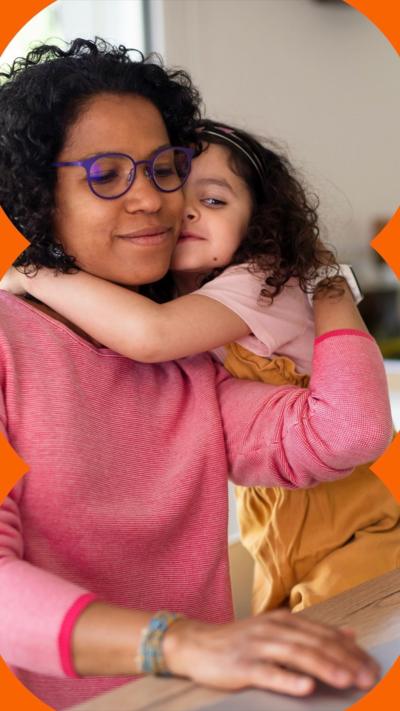 A woman wearing a pink top hugs a girl in a pink top and yellow dress. They both have curly hair.