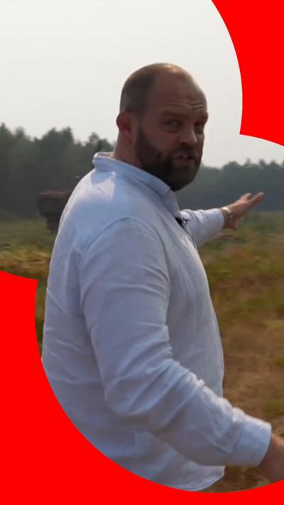 BBC correspondent James Waterhouse wearing white shirt and pointing to forest in the background