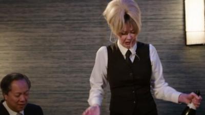Mandy, in a waitress outfit, holds an open bottle of champagne while looking stressed