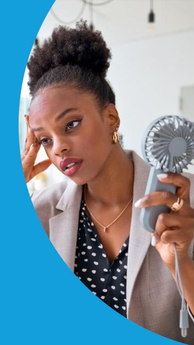 A woman sat in front of a computer screen, holding a mini fan, looking tired