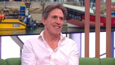 Rob Brydon in a pink shirt sitting on a green sofa smiling