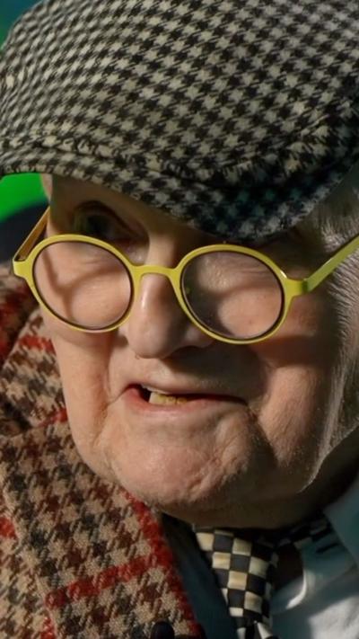 David Hockney wearing a flat cap and round yellow glasses is looking into a camera and talking.