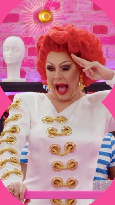 Drag queen La Voix salutes. She has curly red hair and wears a nautical themed white dress