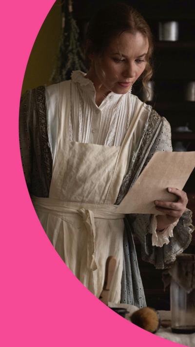 An actress playing Jane Austen. They are in a room lit by candle light and reading from a piece of paper.