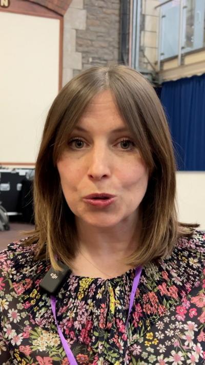 Woman with brown bob speaking into the camera, wearing a floral dress