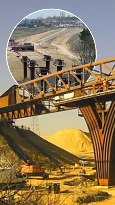 Bridge slowly being put into place on a sunny day, workers and vans can be seen below it.