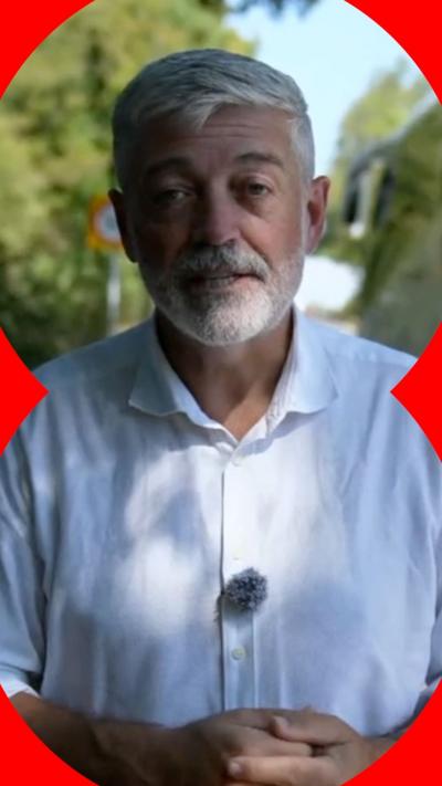 A man with short grey hair, beard and moustache is standing in a shady spot. He is wearing a white shirt.