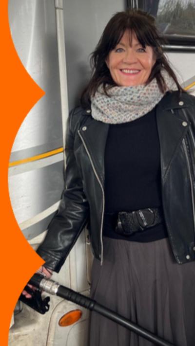 A woman with long dark hair, wearing a white scarf and a leather jacket fills up the fuel of the camper van.